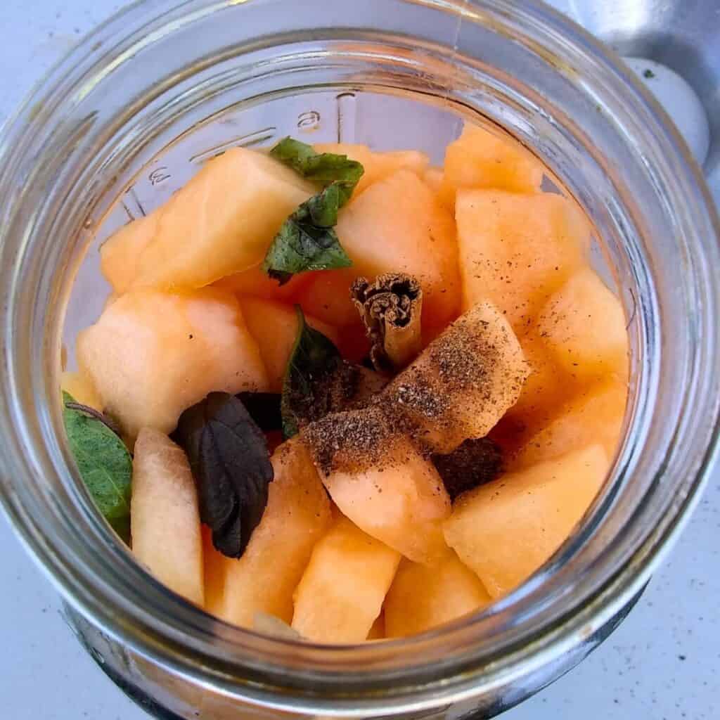 a mason jar full of cantaloupe pieces with spices and basil leaves, viewed from above.