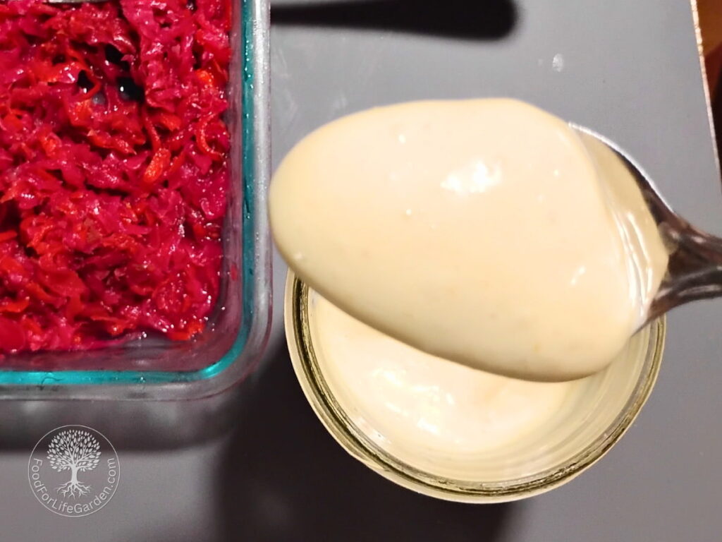 A spoon full of fermented mayo in the forground. below is a jar filled with mayonnaaise and next to it a dish with fermented coleslaw.