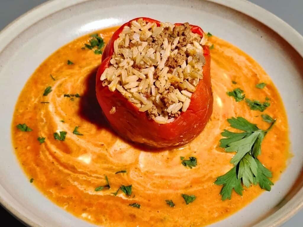 a plate with a red bell pepper stuffed with beef and rice filling, placed in a sea of orange and red garden vegetable gravy with white swirls of sour cream. decorated with sprinkles and a sprig of parsley.