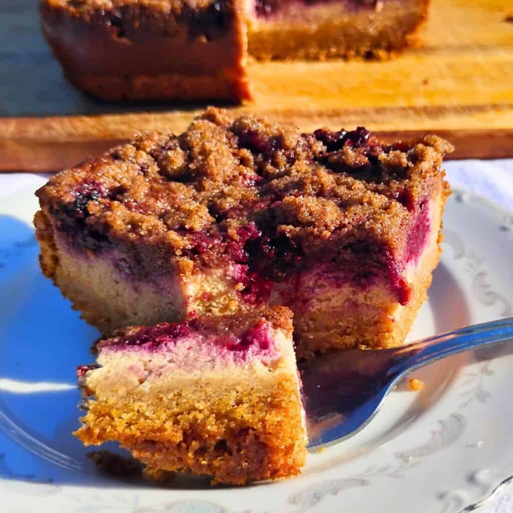 a slice of blackberry cheesecake streusel bars on a plate with a fork having removed a piece. In the background there is a whole cake with a piece removed.