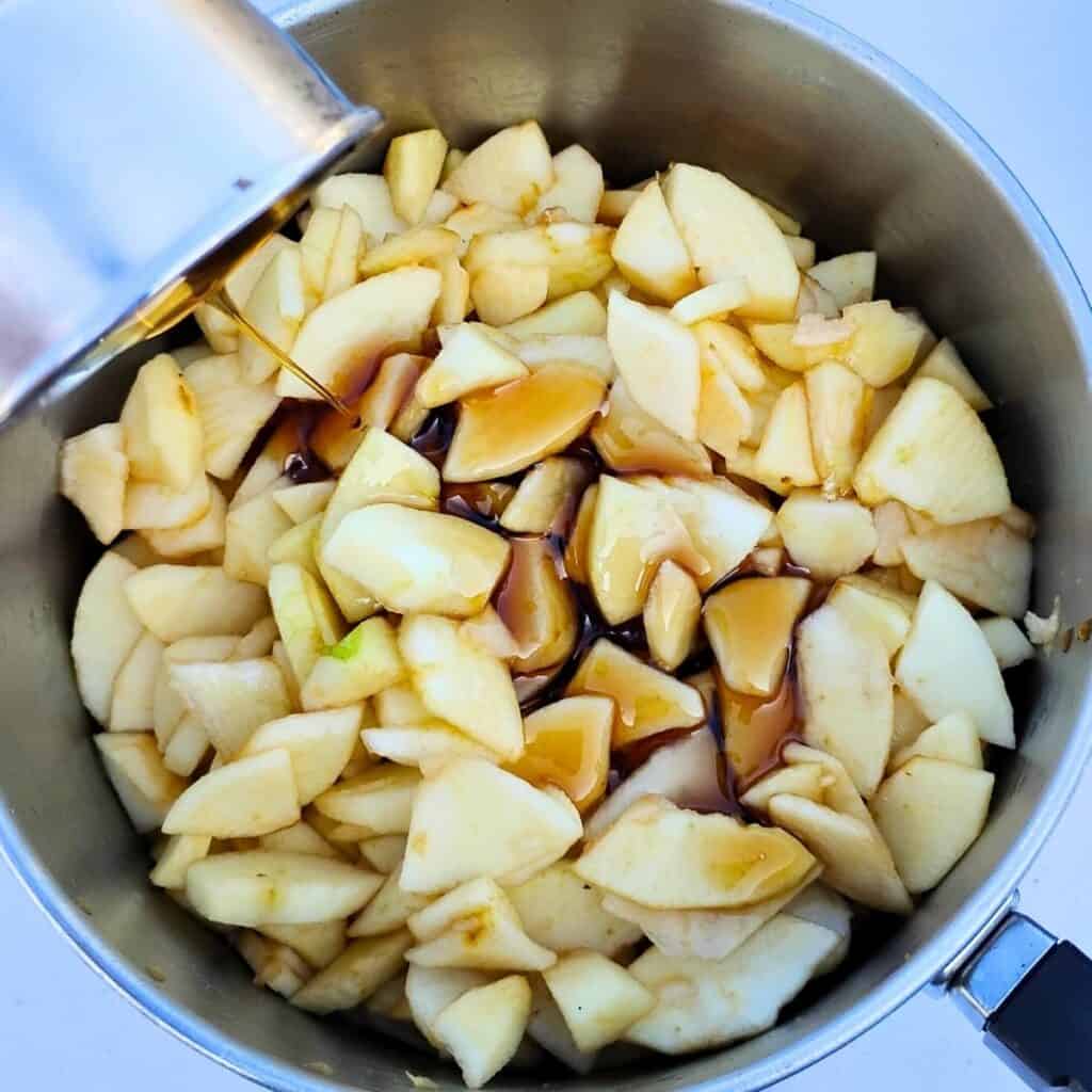 apples in a pot are getting maple syrup poured over them.