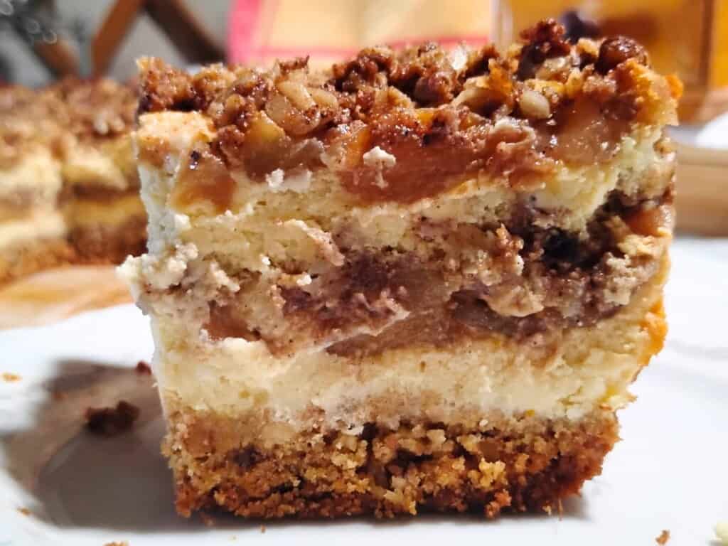 A piece of apple pie cheesecake on a plate with the whole cake behind it.