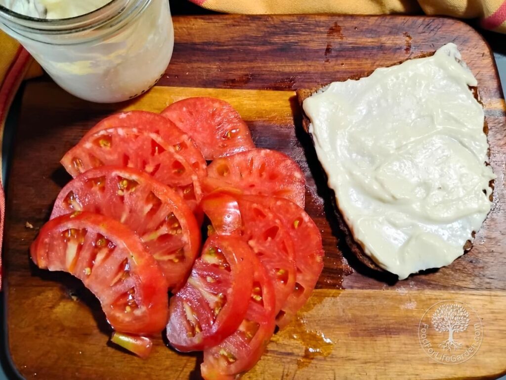 a cutting board with a piece of kamut and einkorn bread topped with mayonnaise. beside it are slices of tomato, and a jar filled with homemade mayonnaise.
