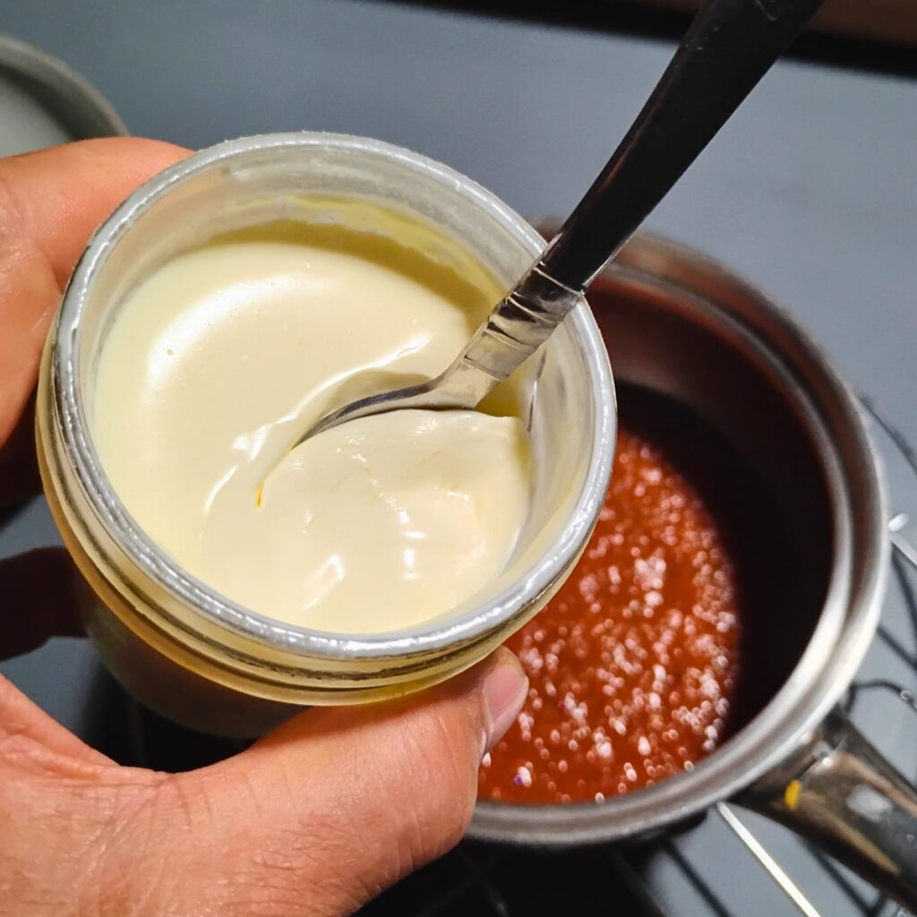 photo of a hand holding sour cream in a jar with a spoon in it. there is a pot with vegetable sauce below.