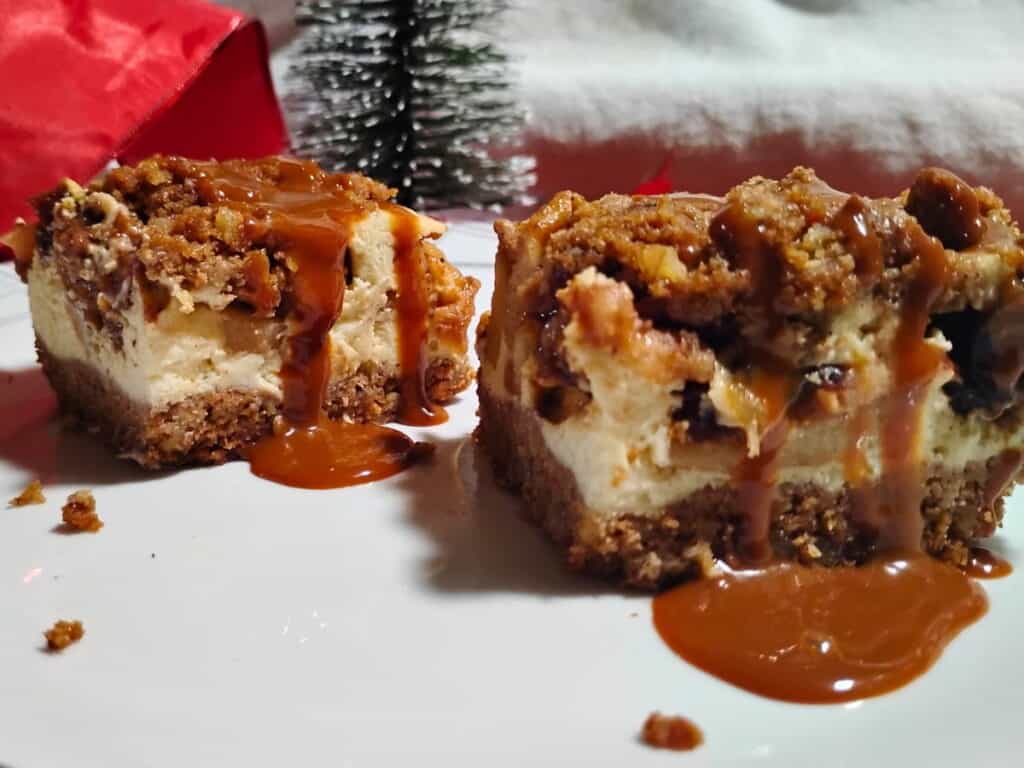 A festive holiday setting with two caramel apple pie cheesecake bars on a plate. There is a Christmas tree decoration behind it and some red christmas ribbon.