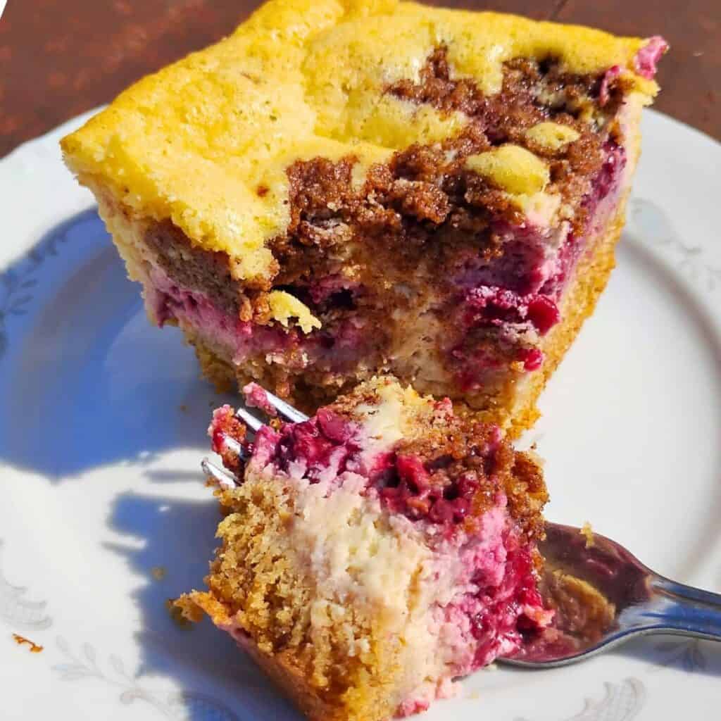 a piece of blackberry cheesecake streusel bar on a plate with a fork that has a small piece on it to show off the texture.