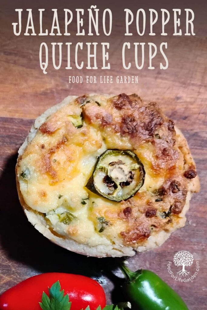 photo shows a mini jalapeno popper quiche cup on a wooden board with a slice of jalapeno decorating it. there is a red and a green jalapeno in front of it. text overlay reads jalapeno popper quiche cups. food for life garden. 