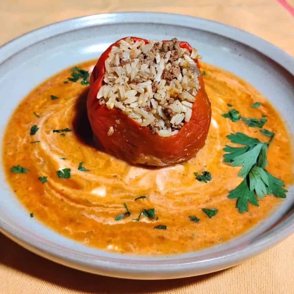 photo shows a plate with a red healthy stuffed pepper filled with beef and rice in a puddle of red and orange vegetable sauce with white swirls of sour cream and a sprinkle of parsley.