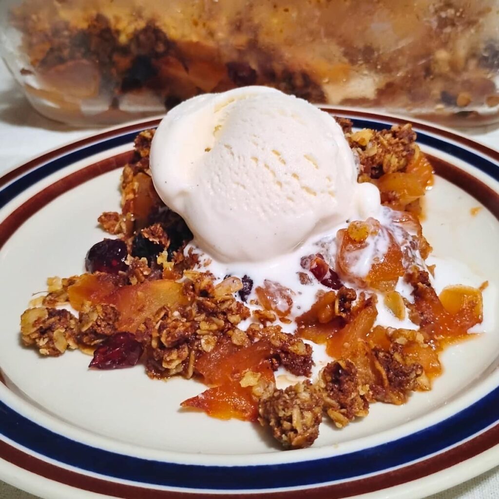 A plate with some healthy apple crisp and a scoop of ice cream on top. there is a baking dish with more apple crisp behind it.