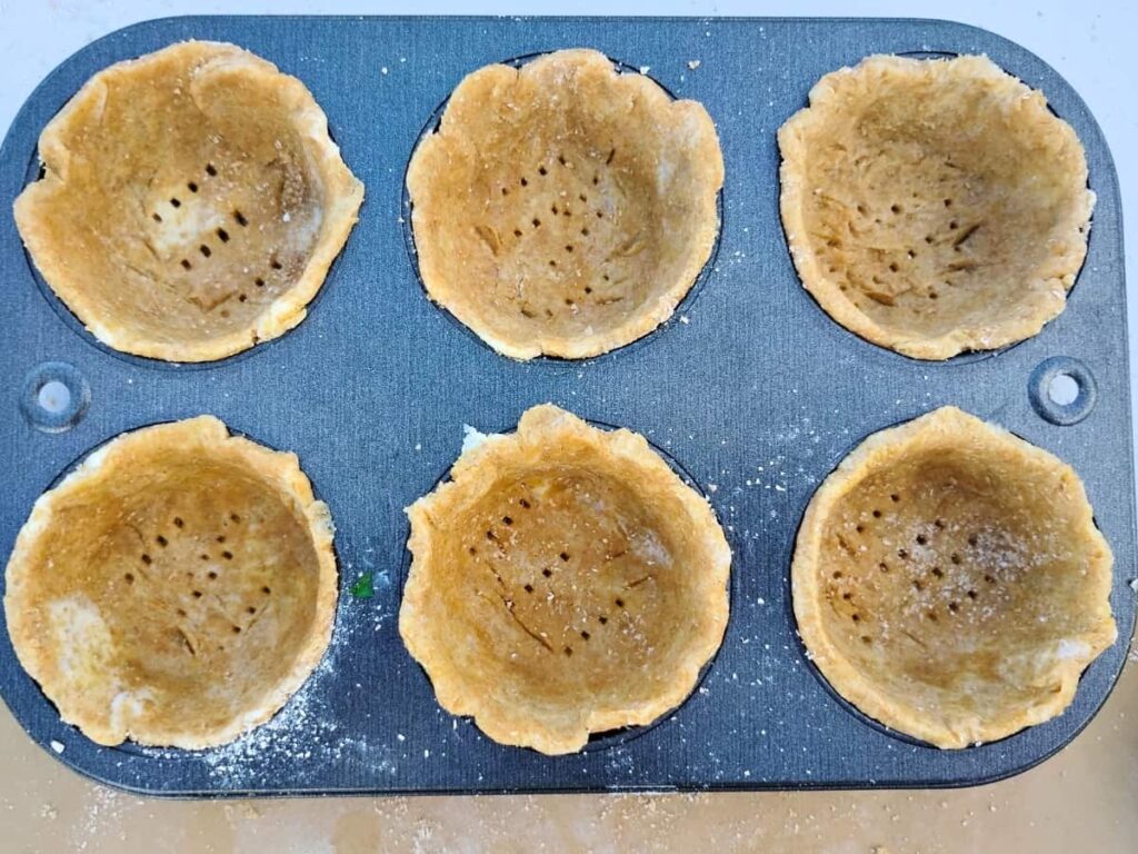 A muffin tin holding 6 empty flaky pie crust cups that have been pierced with a fork at the bottom. 