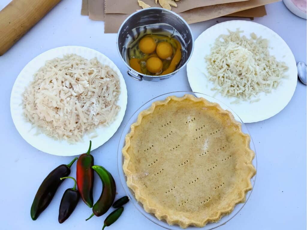 ingredients on a table for jalapeno pie. two varieties of grated cheeses, eggs in a bowl, and jalapenos.