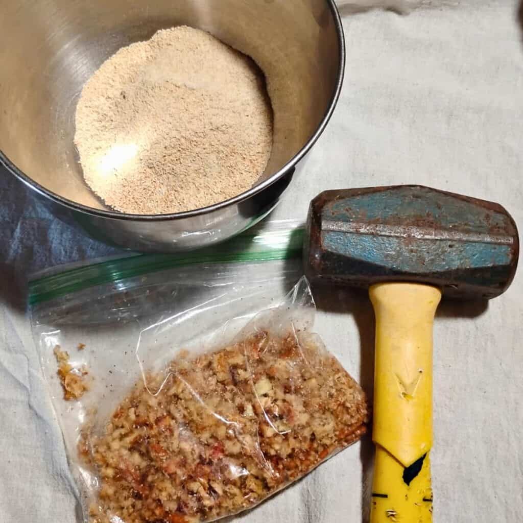 a bowl with flour and a plastic baggie with smashed up pecan pieces with a hammer next to it. 