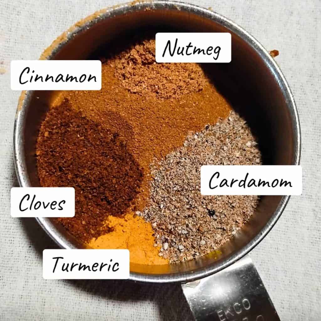 measuring cup with measured out spices, nutmeg, cinnamon, cloves. turmeric and cardamom. 