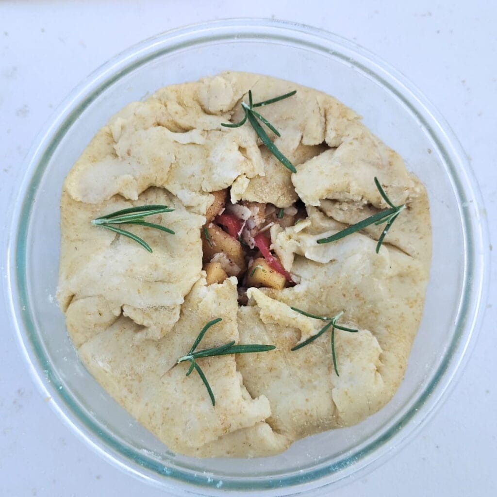 a small rustic style apple pie in a glass dish with the edges folded toward the middle. It is decorated with rosemary sprigs. 