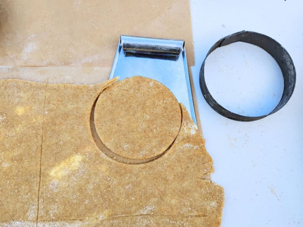 Demonstration of cutting the pie crust with a circular cutter and using a pastry cutter to lift it off the parchment below.