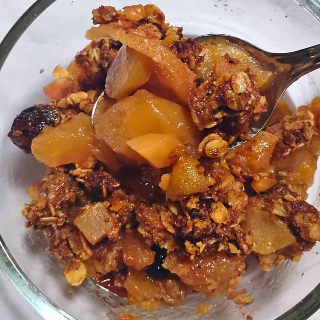 a bowl filled with ginger spice healthy apple crisp with cranberries and a oat and walnut crumble topping. A spoon is removing a portion of it.