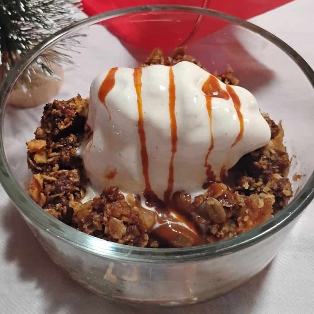a bowl filled with ginger and cranberry apple crisp and a scoop of ice cream with goat milk caramel, cajeta, drizzled over it. There is some holiday decoration in the background.