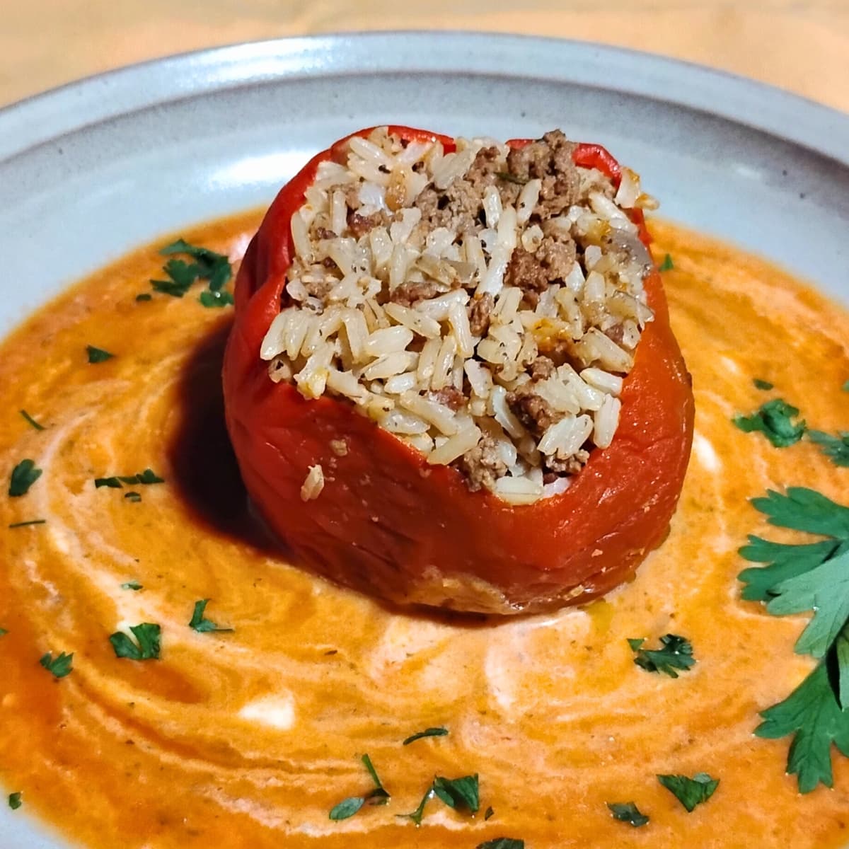 photo shows a plate with a red healthy stuffed pepper filled with beef and rice in a puddle of red and orange vegetable sauce with white swirls of sour cream and a sprinkle of parsley.