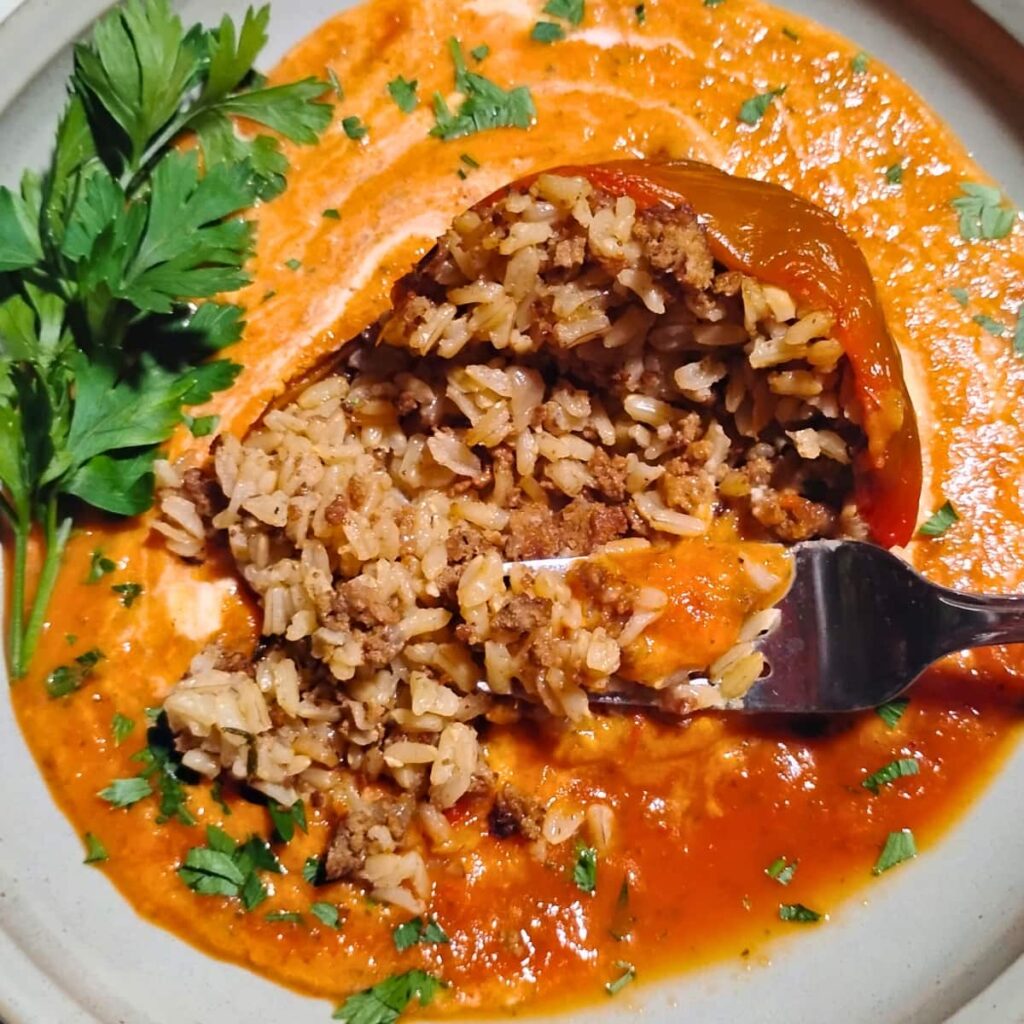 photo shows a fork digging into a red bell pepper stuffed with rice and beef filling, laying in a puddle of red and orange vegetable sauce with white sour cream swirls and a sprinkle of parsley.