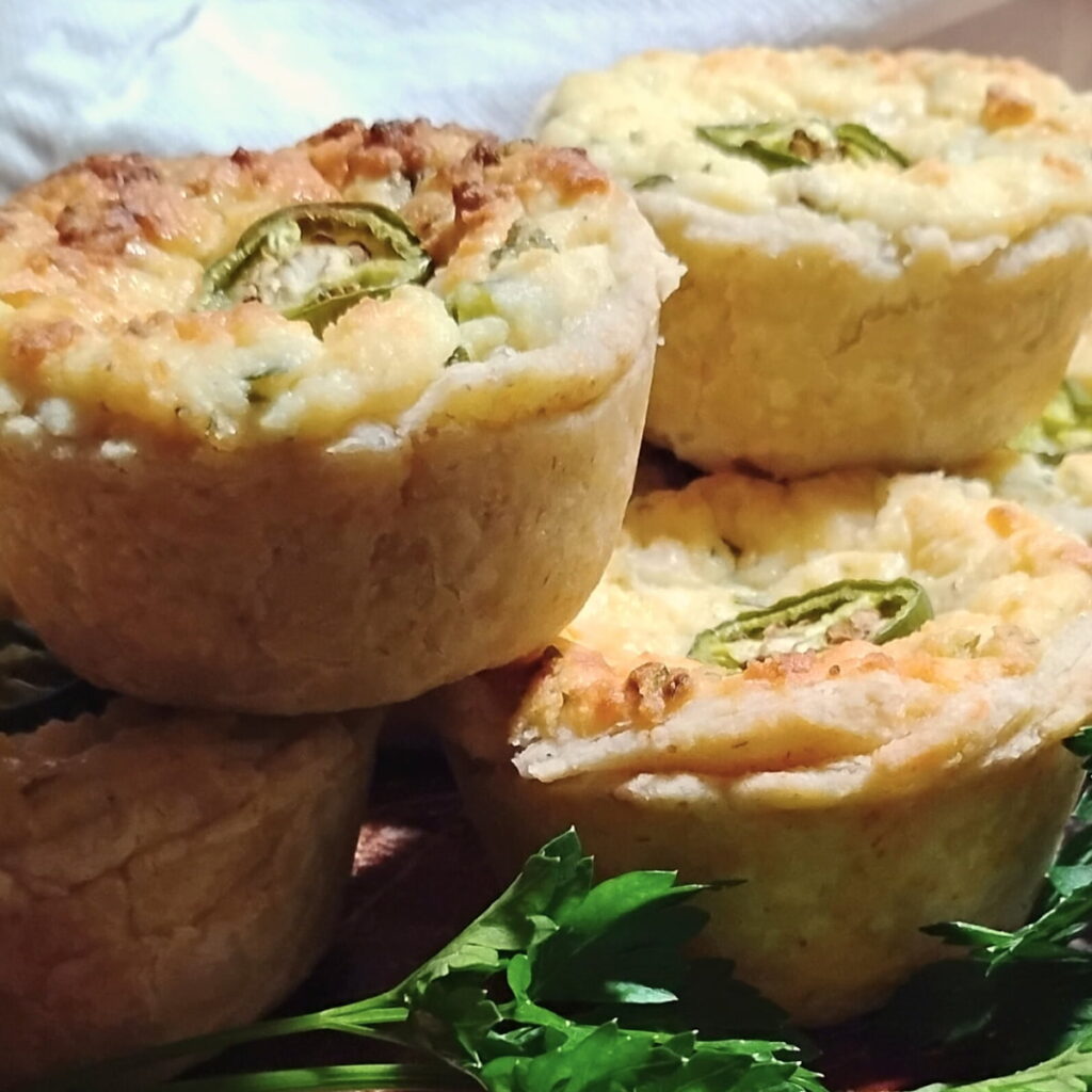 A pile of mini jalapeno popper quiche cups on a baking sheet, decorated with sliced jalapeno pieces. There is some parsley in the foreground. 