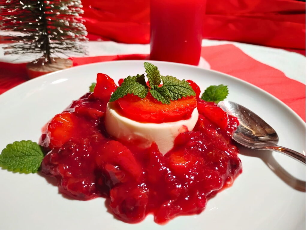photo of a single serve portion of panna cotta with some raspberries on top and surrounded with cranberry orange and hibiscus sauce with lemon balm leaves for decoration. there is a candle and christmas tree behind it.