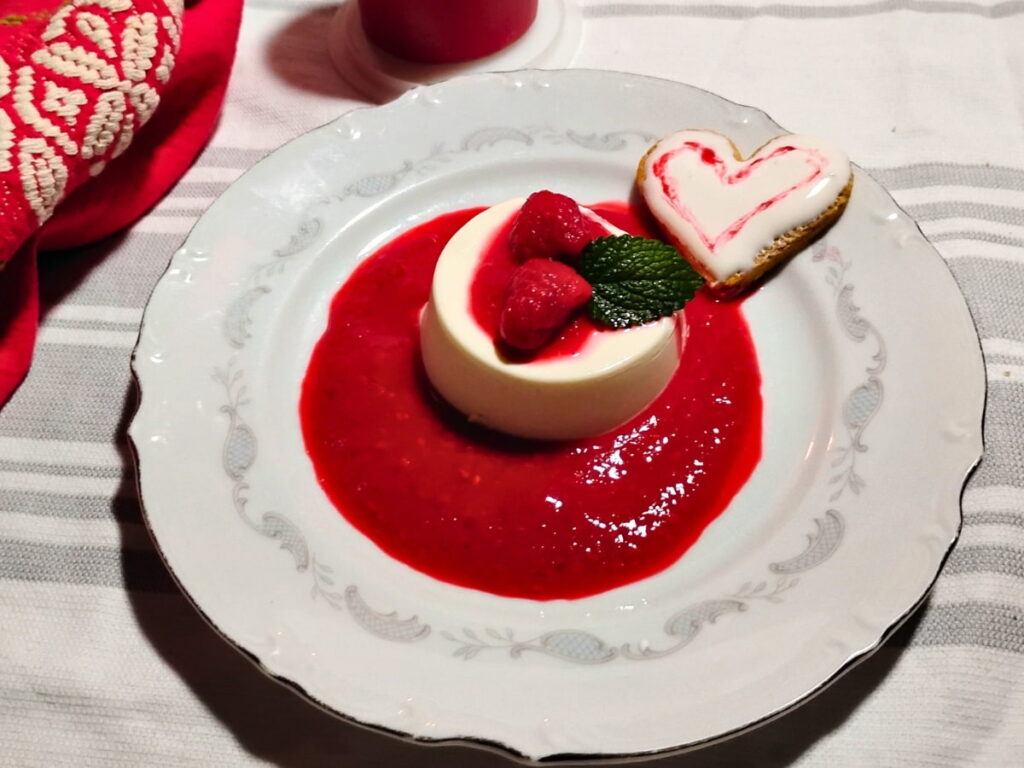 photo of a single serve portion of probiotic panna cotta decorated for valentine's day with some raspberries on top and surrounded with raspberry sauce. there is a small heart shaped cookie with frosting beside it.