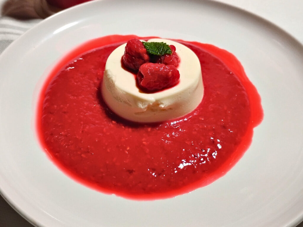 photo of a single serve portion of panna cotta with some raspberries on top and surrounded with raspberry sauce.