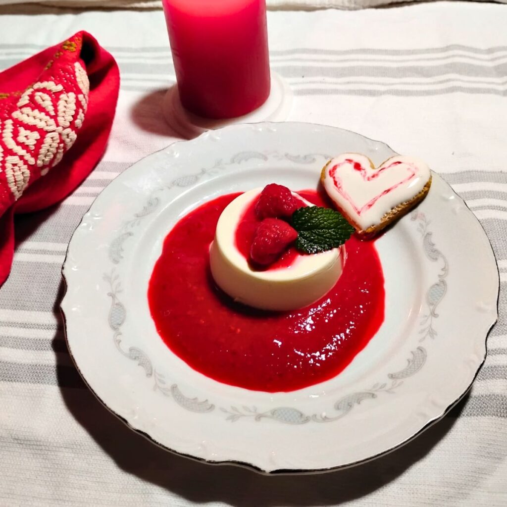 photo of a single serve portion of panna cotta with some raspberries on top and surrounded with raspberry sauce. There is a heart shaped cookie with frosting on the plate too and a candle and napkin behind it.