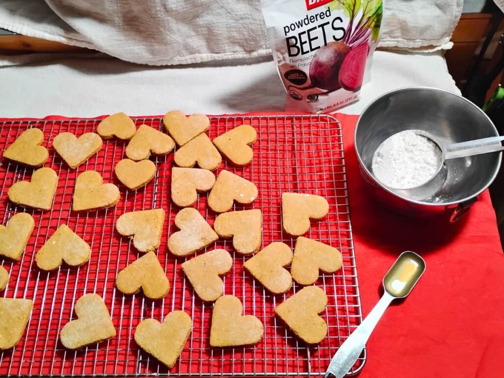 a cooling rack with lemon heart cookies and ingredients for making the icing. Lemon juice, powdered sugar, and beet powder.