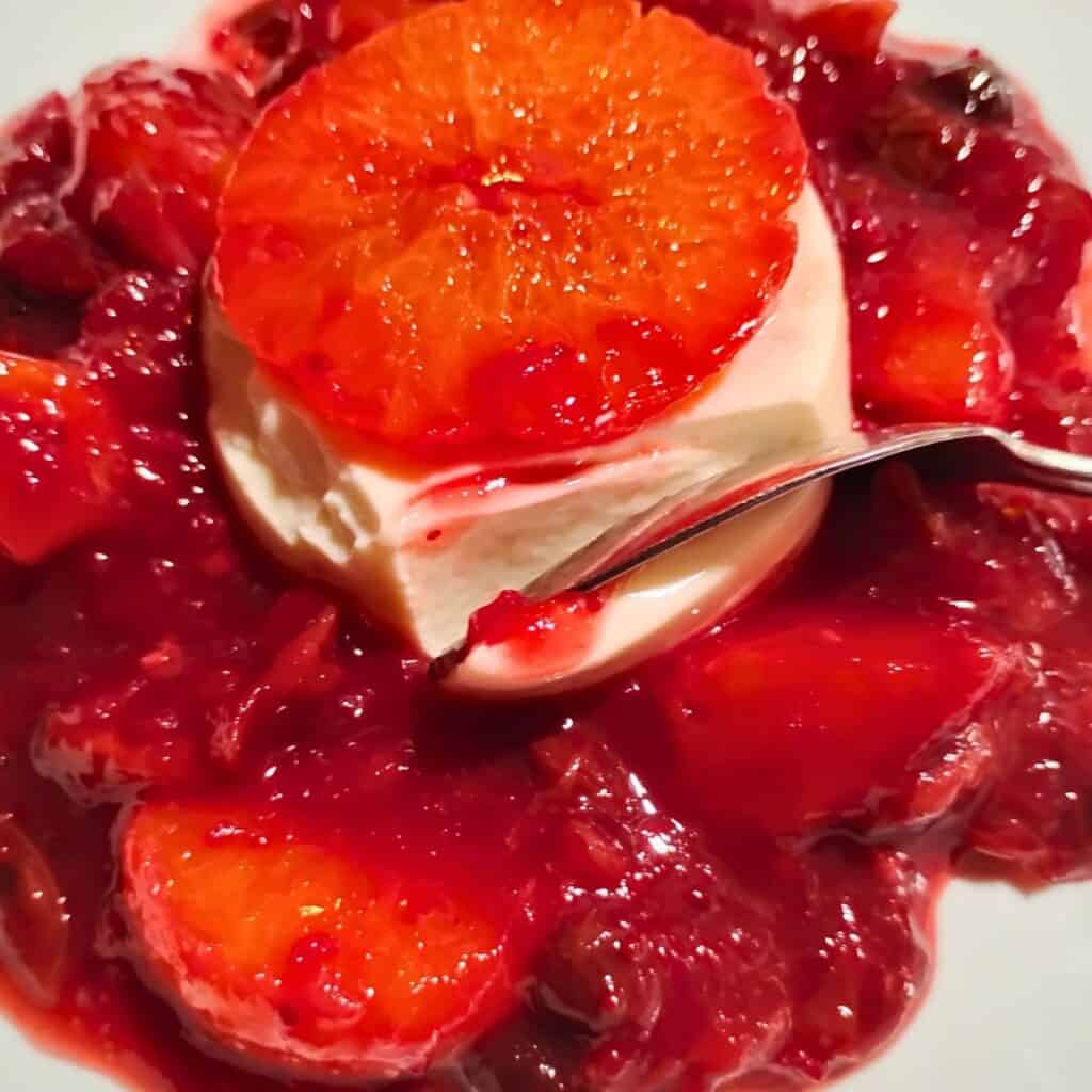 photo of a bed of cranberry orange sauce with a small molded panna cotta in the middle of it. A slice of mandarin is on the top. and a spoon is removing some of the panna cotta.