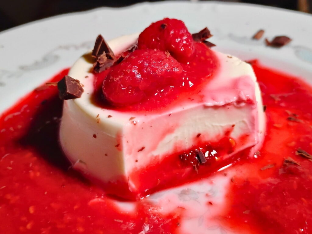 photo of a single serve portion of panna cotta with some raspberries on top and surrounded with raspberry sauce. A portion of the panna cotta is removed, showing off the awesome creamy inside.