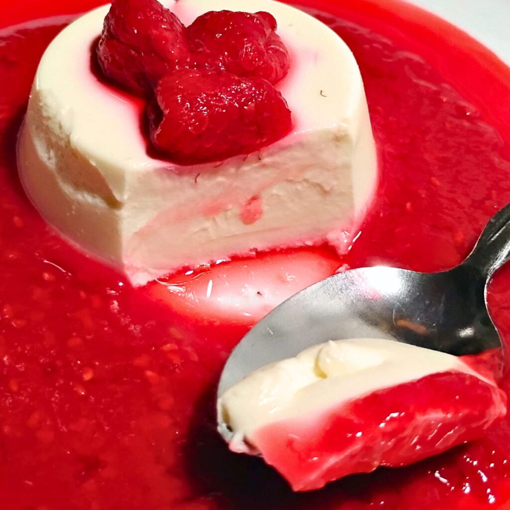 a single serve portion of probiotic panna cotta with some raspberries on top and surrounded with raspberry sauce. A portion was removed with a spoon and is leaving a view of the creamy texture of the panna cotta.