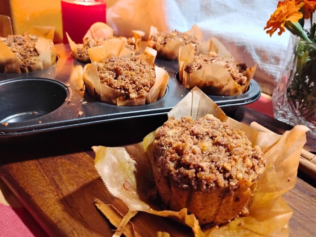 photo of a muffin inside a liner on a cutting board with a muffin pan filled with muffins behind it. there are some fall marigolds in a glass and a lit candle.