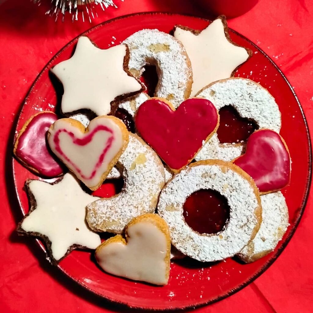 A Christmas cookie platter with a mix of cookies piled up on it. zitronenherzen, vanillekipferl, zimtsterne and linzer cookies.