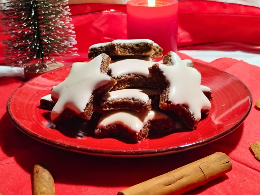 A pile of zimtsterne cinnamon stars on a red plate with a candle and fake christmas tree decoration behind it and cinnamon sticks on a red napkin in front of it.