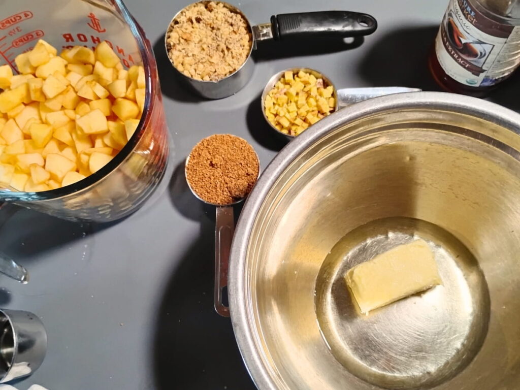 Some ingredients are shown for this recipe. Chopped apples, chopped nuts, chopped ginger and panela sugar in measuring cups. butter and oil in a bowl, and vanilla extract in a bottle.