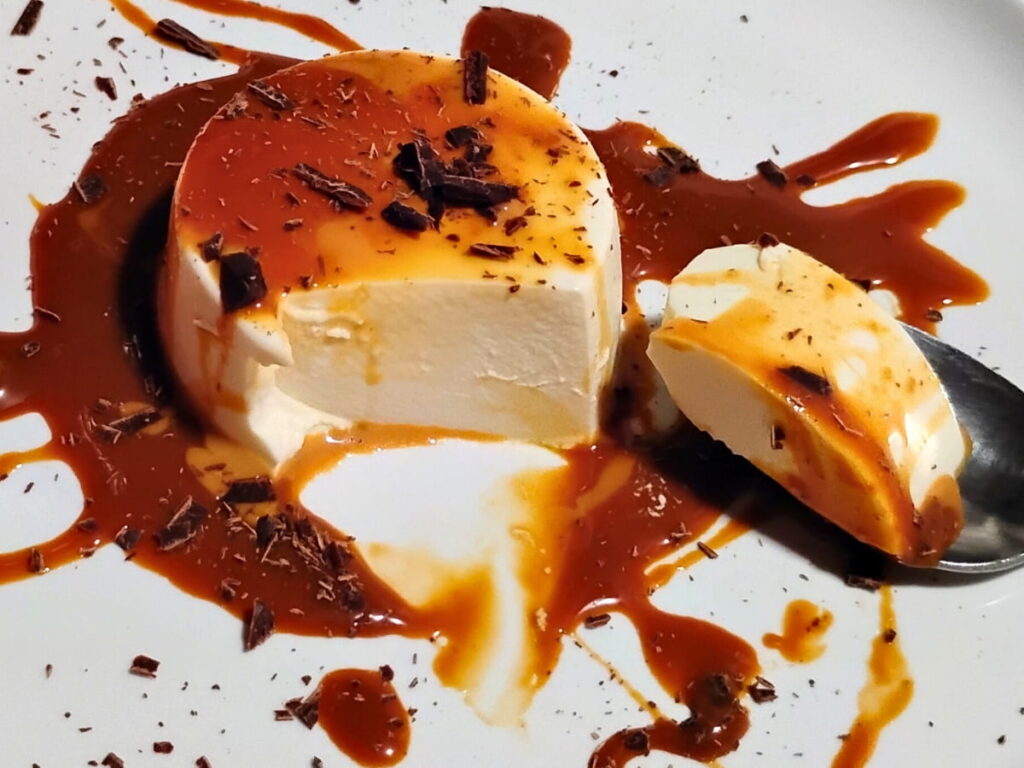 photo of a single serve portion of panna cotta with some raspberries on top and covered and surrounded with cajeta caramel sauce. A spoon has removed a portion and the creamy texture of the inside is showing.
