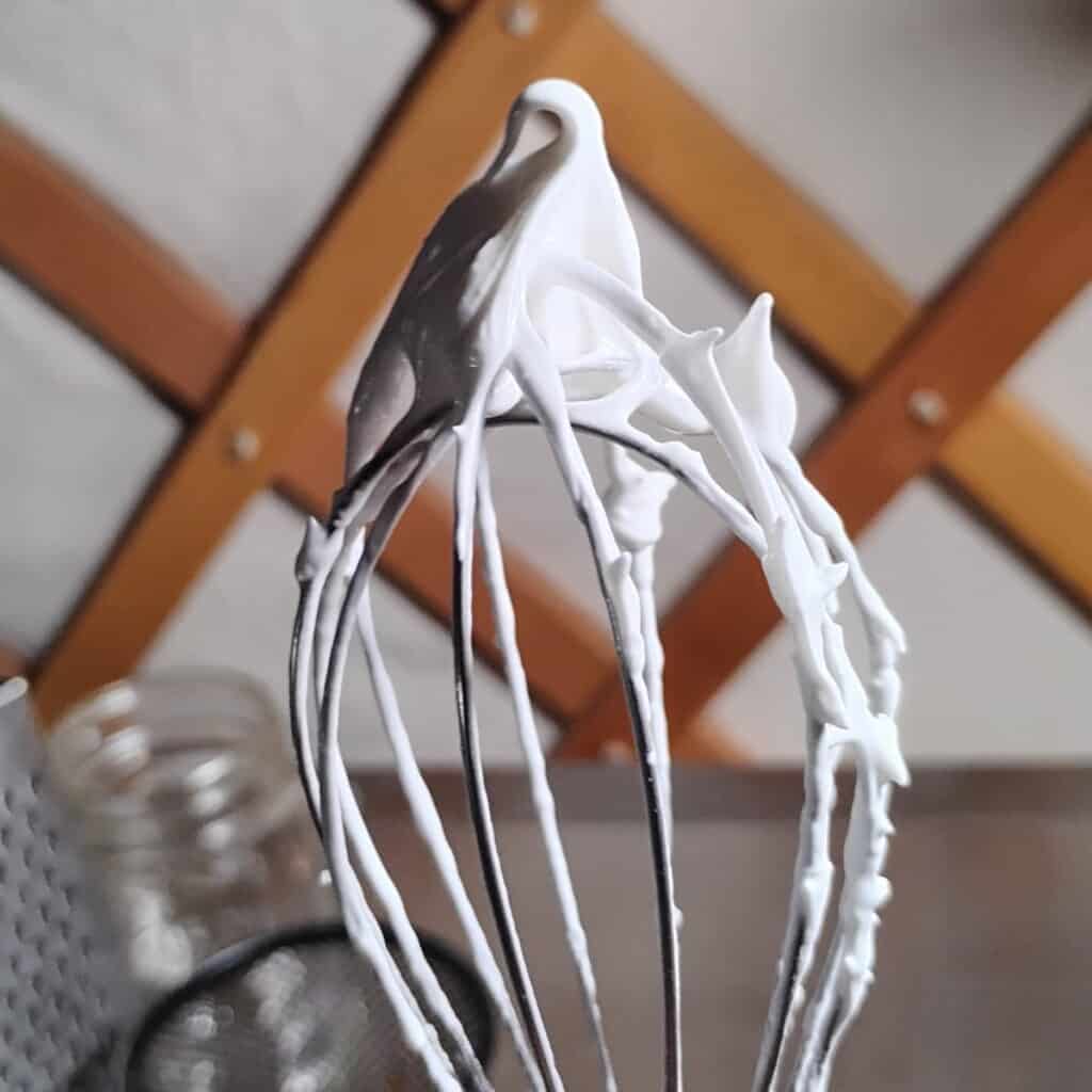a whipping whisk with a tuft of meringue fluff with a curly cue at the top, showing the perfect consistency of the meringue batter.