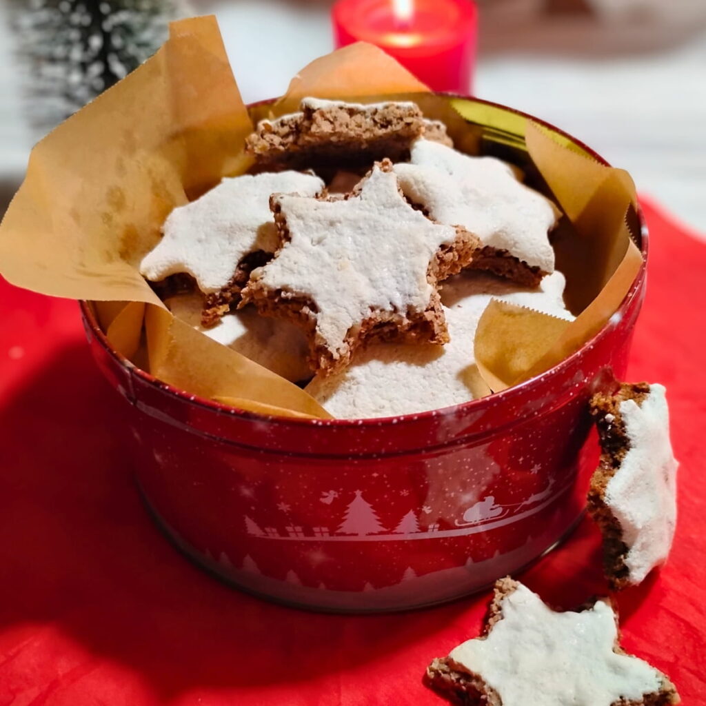Christmas cookie tin filled with zimtsterne cinnamon stars and a few are leaning against it on the outside. there is a candle and decorative christmas tree behind it.