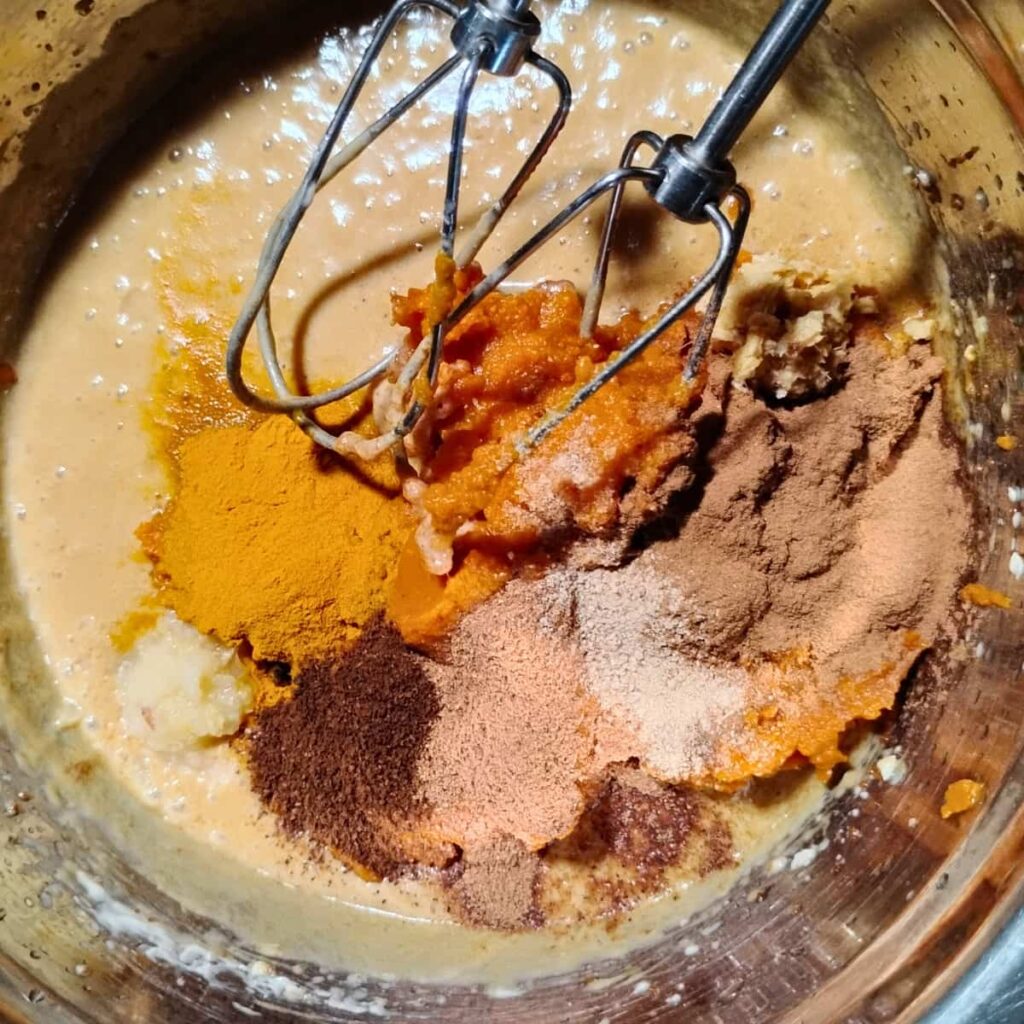 a bowl with batter getting prepared and pumpkin puree and spices dumped in. two beaters are shown.