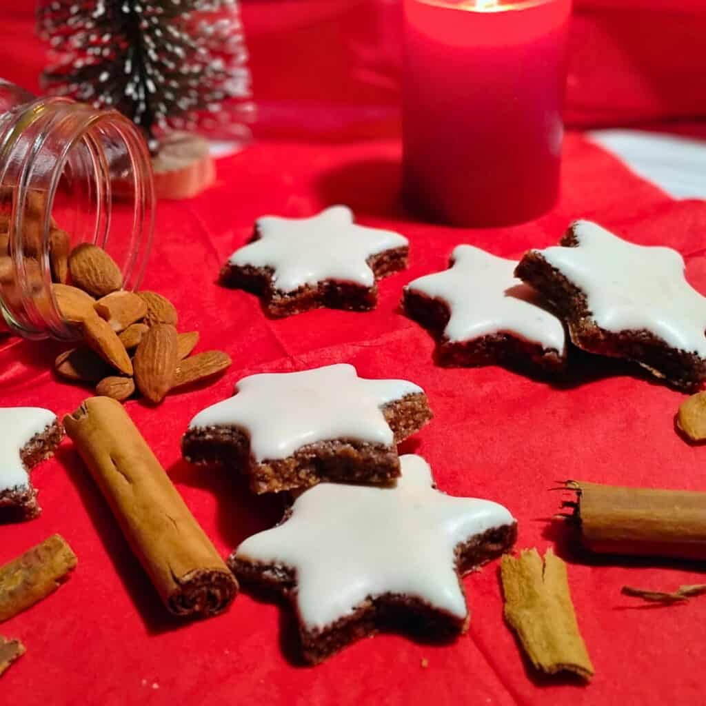 Zimtsterne or Cinnamon Stars are scattered on a red napkin mixed with cinnamon sticks and some nuts are spilling out of a glass jar beside it. there is a candle and a fake christmas tree decoration in the background.