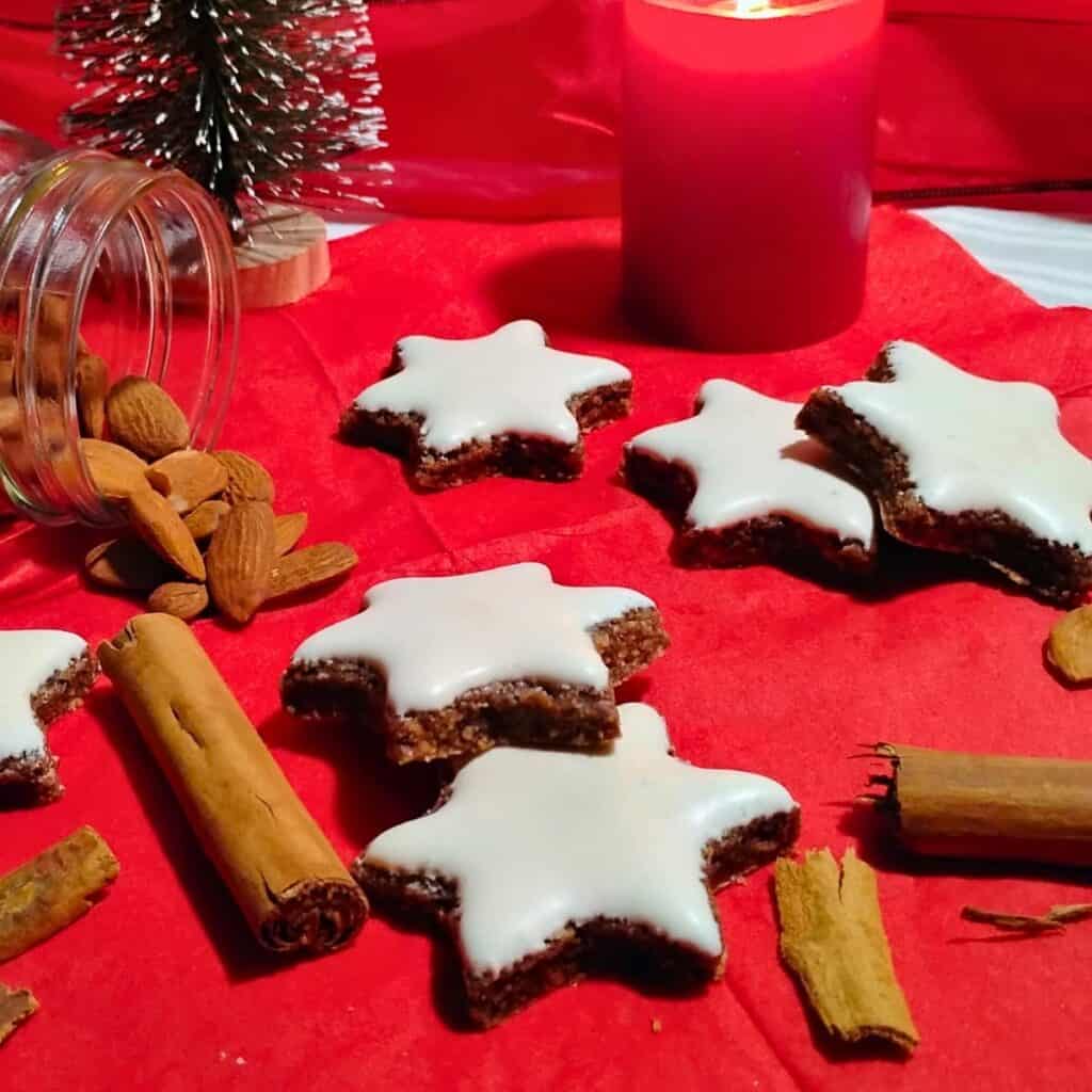 Cinnamon stars on a napkin with a candle and a decoratie christmas tree behind it. There are spices and a spilled jar with almonds.