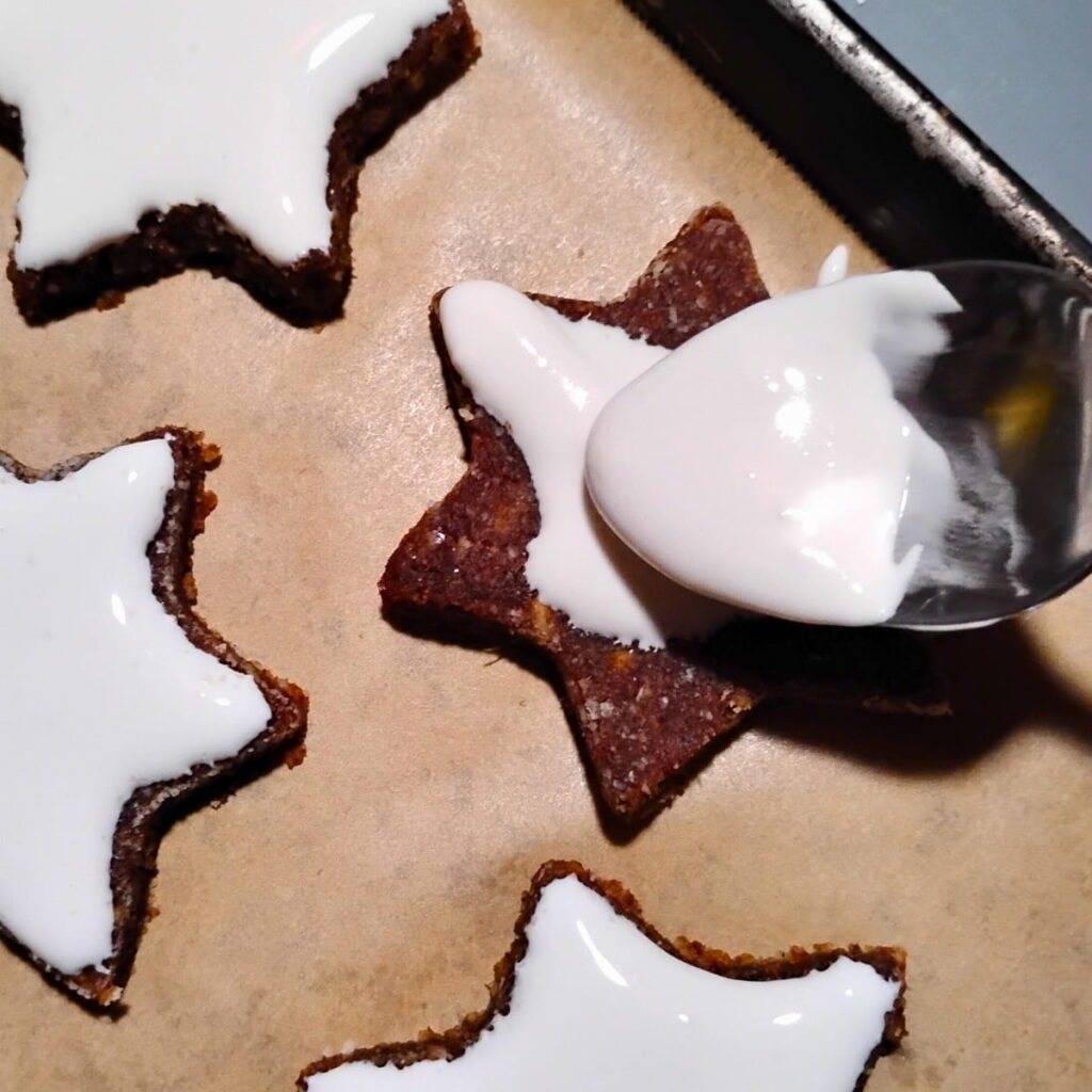 Cinnamon stars on a baking sheet are getting frosted using a spoon with white fluff frosting.
