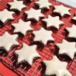 Frosted German zimtsterne or cinnamon stars lined up on a cooling rack with a red background.