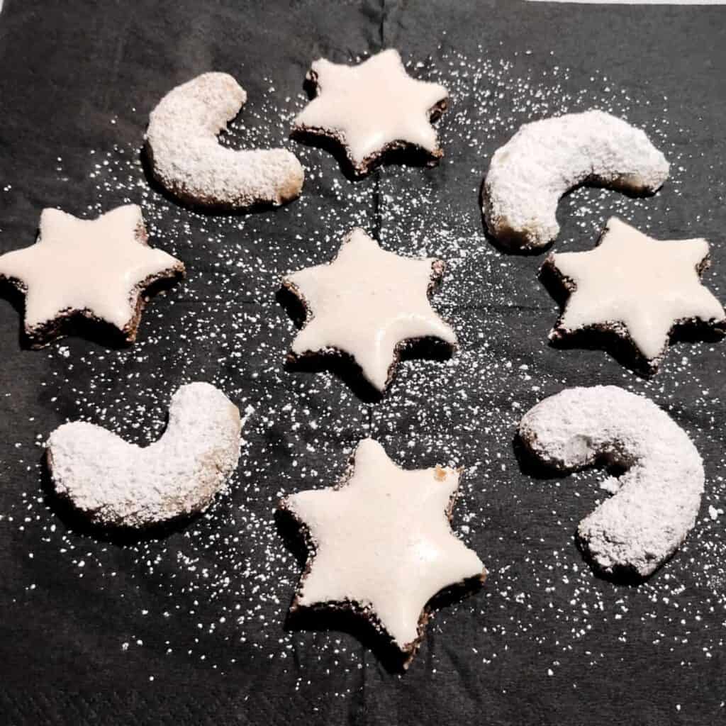a few Zimtsterne cinnamon stars and almond Vanillekipferl arranged on a black napkin with some powder sugar sprinkeled over it.
