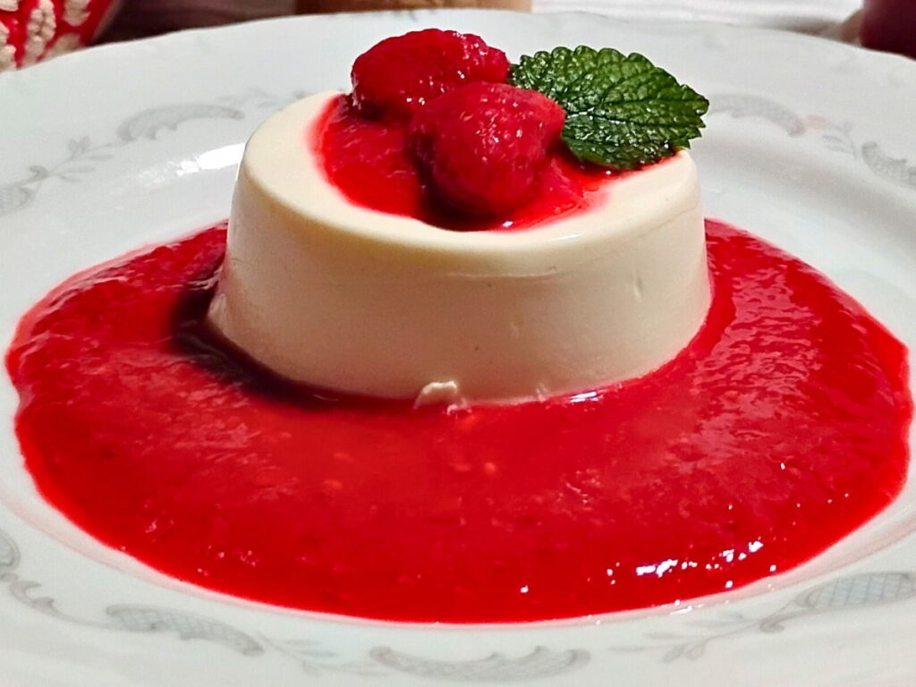 a plate with a portion of probiotic panna cotta sitting in a sea of fresh raspberry sauce with some raspberries on top and a leaf of lemon balm.