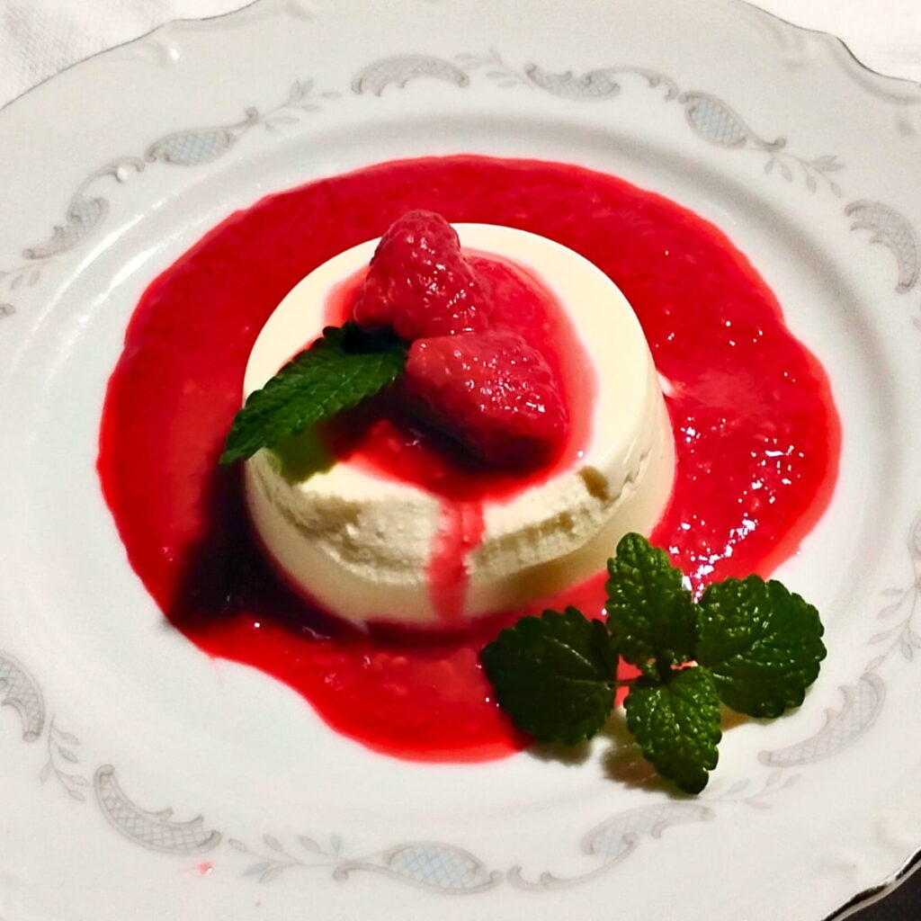 photo of a single serve portion of probioticpanna cotta with some raspberries on top and surrounded with raspberry sauce and decorated with lemon balm leaves on a white plate.