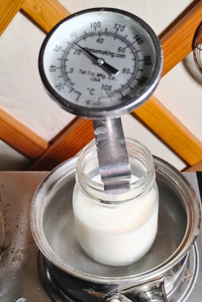 milk is getting heated in a pint jar with a thermometer in it, sitting in a water bath in a small pot. 