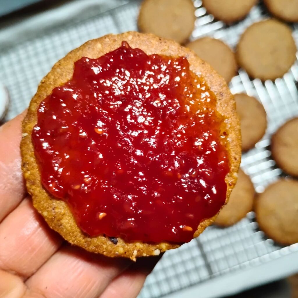 close-up photo of a cookie bottom in a persons hand with jam spread over it. More cookies to be filled are below on a rack.