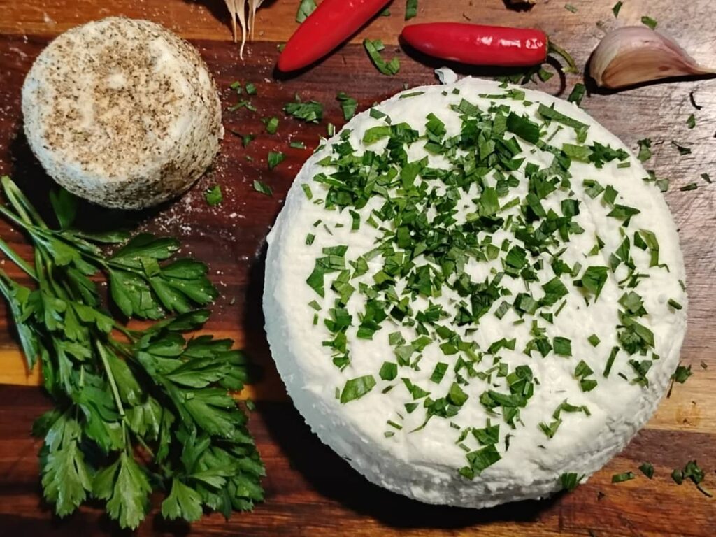 formed chevre cheese on a wooden cutting board. one is rolled in black pepper, the other sprinkled with parsley. there are hot peppers and garlic on the board too.